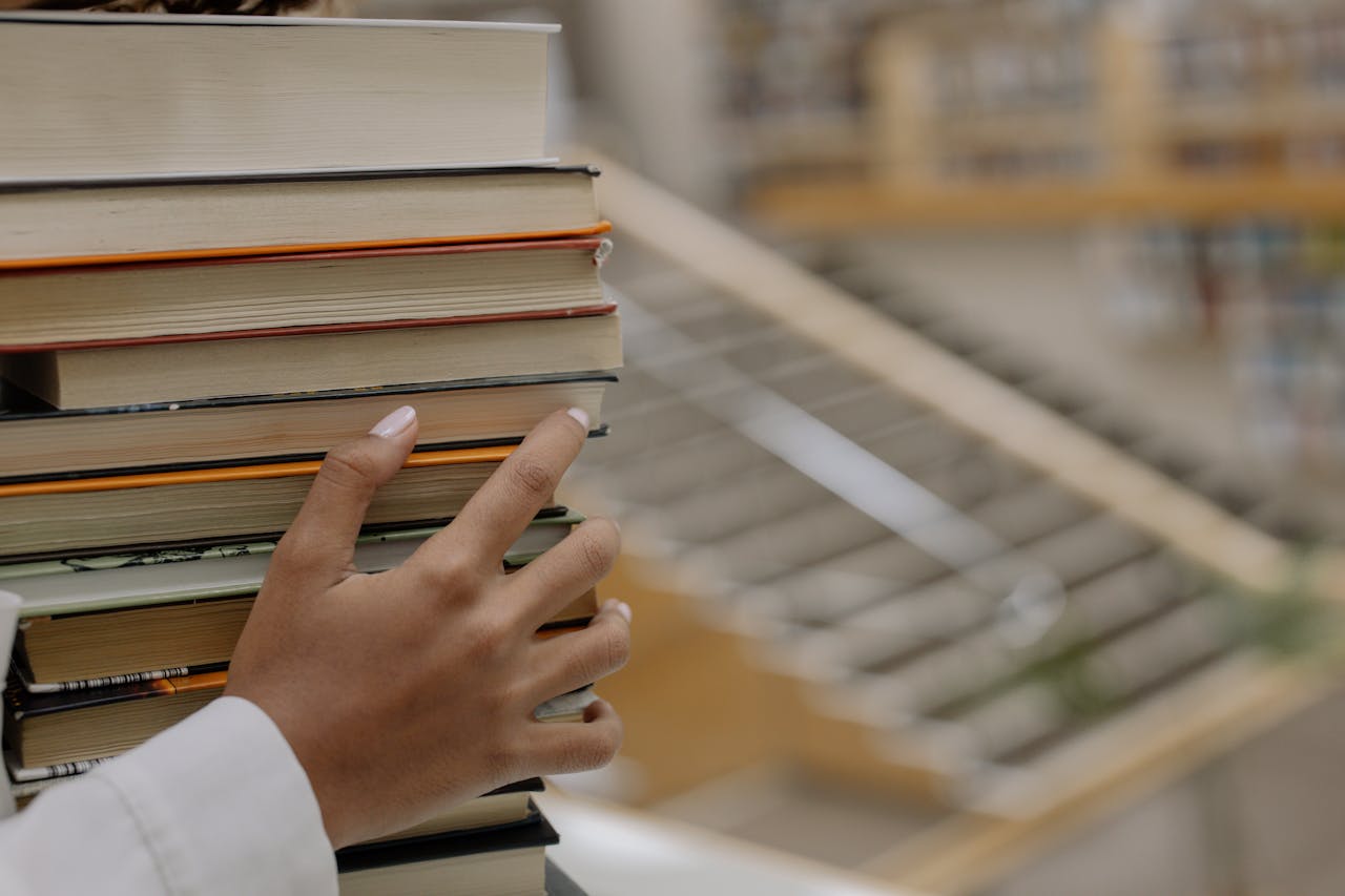 A hand holding a stack of books with bookshelves blurred in the background, depicting knowledge and learning.